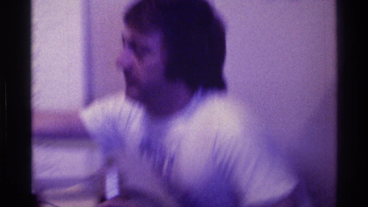Blurred young man in a white T-shirt seated indoors beside a plain wall.
