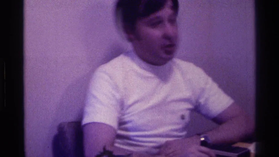 1976 Kentucky home-movie frame showing a young man in a white graphic T-shirt seated at a desk against a plain wall.