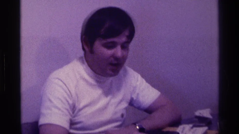 1976 Kentucky 8mm frame of a young man in a white T-shirt seated at a desk with papers visible nearby.