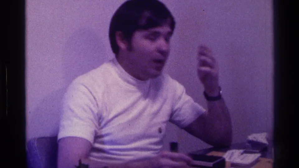 1976 Kentucky home-movie frame of a young man gesturing at a desk, with a small microphone and papers visible.