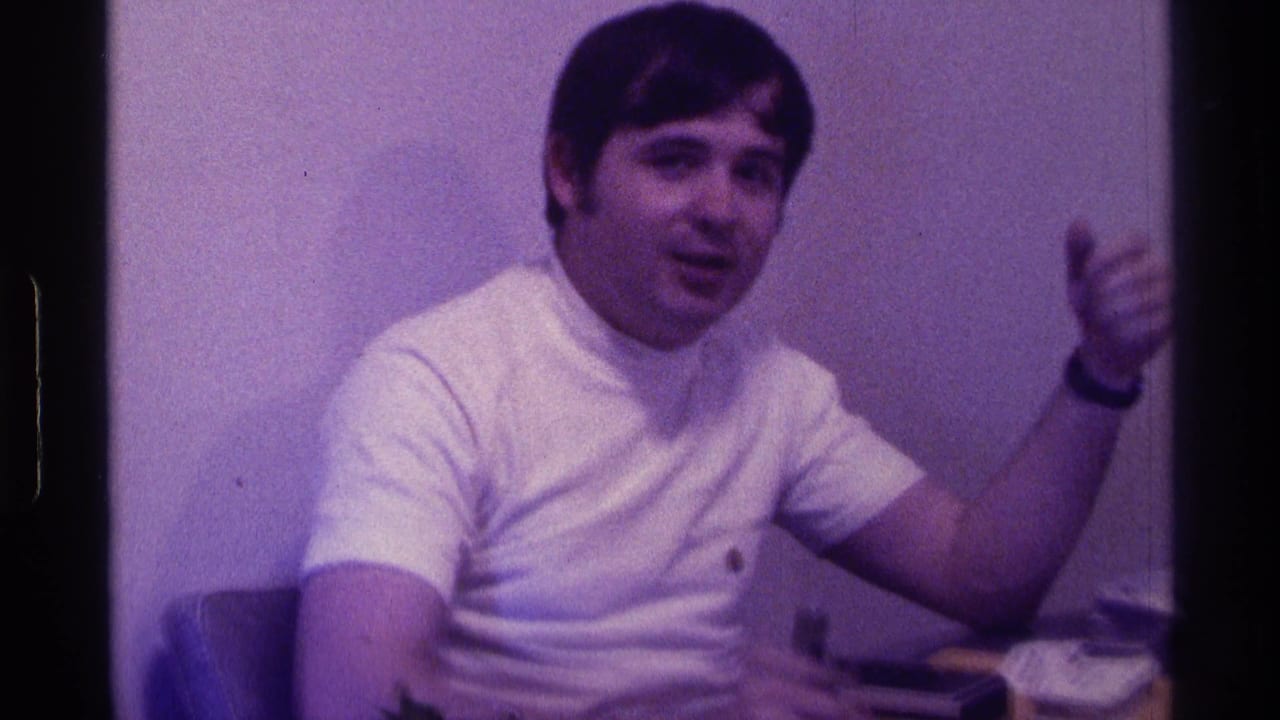 Young man in a white T-shirt sits indoors and gestures toward the camera.