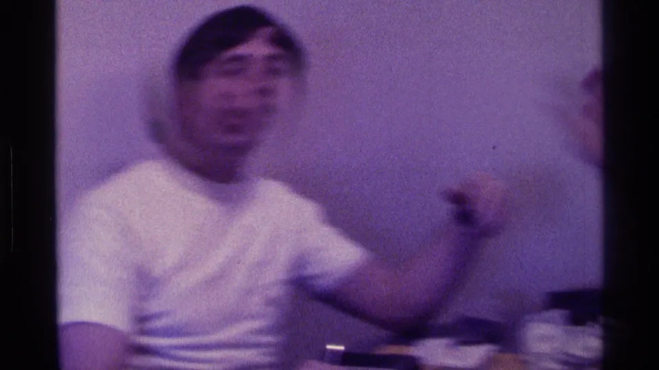 A blurred 1976 Kentucky frame shows a young man in a white T-shirt gesturing animatedly near a table in a private indoor room.