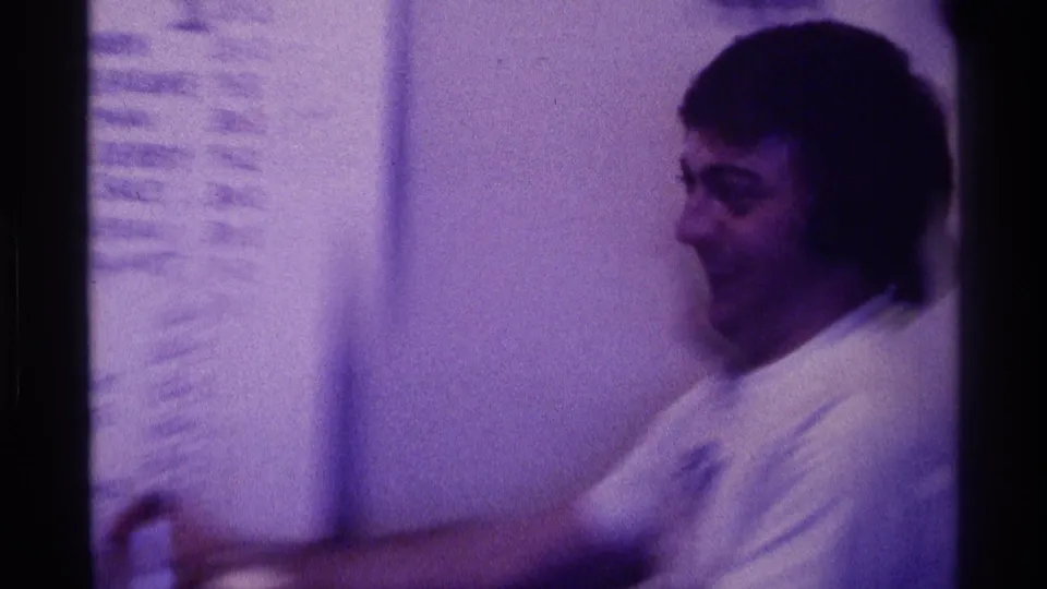 A young man in profile sits near a handwritten board in a Kentucky 1976 home-movie frame, suggesting an informal discussion or game setting.
