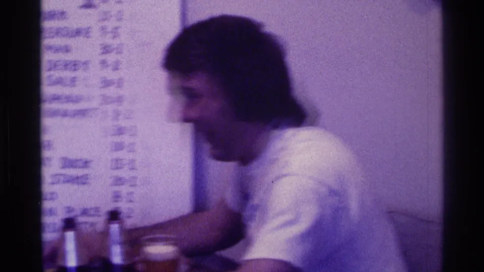 A 1976 Kentucky home-movie frame shows a young man seated at a table with bottles and cups beside a handwritten board.