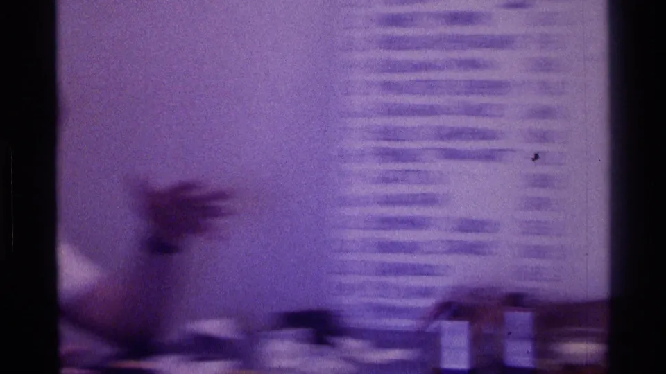 1976 Kentucky 8mm frame showing a blurred seated young man gesturing near table cups and a handwritten list on an interior wall.
