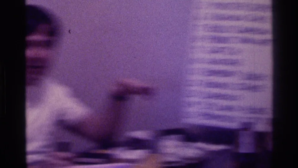 Purple-tinted 8mm frame of a young man in motion beside a table and wall list during a casual indoor gathering in Kentucky.