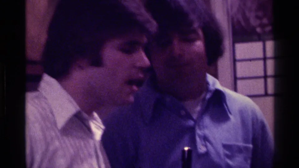 1976 Kentucky home-movie frame showing two young men leaning close in animated conversation inside a private room.