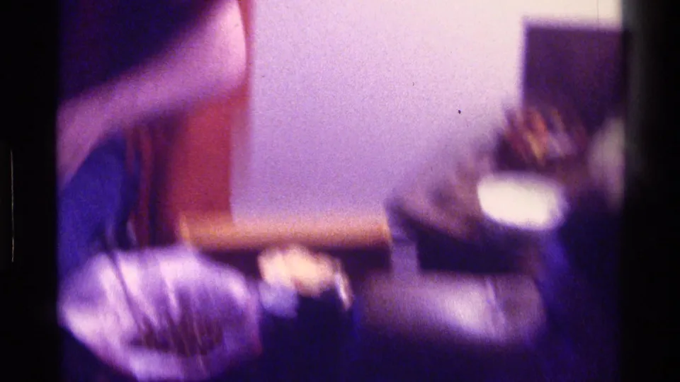 Dim 1976 Kentucky home-movie frame showing a blurred person near upholstered furniture and a small cup-like object.