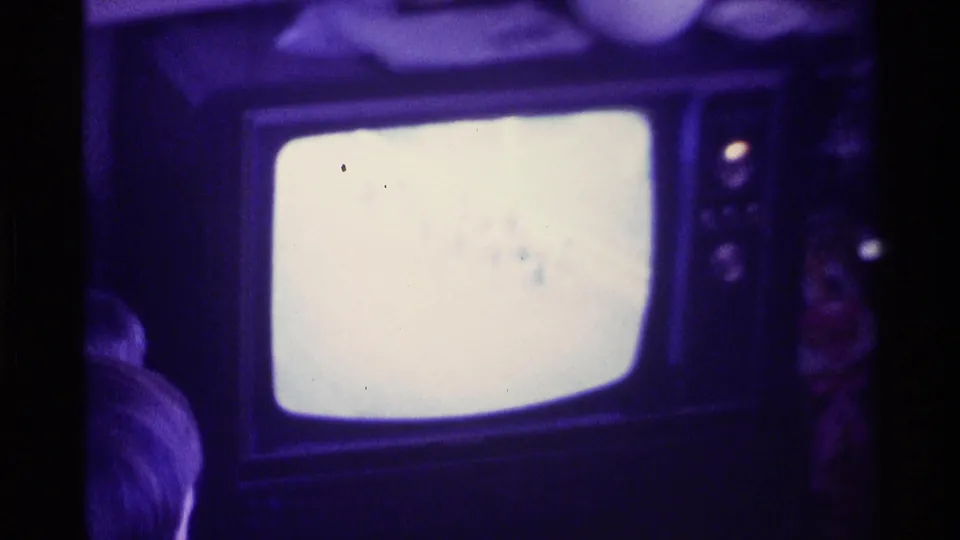 Kentucky 1976 8mm frame showing a glowing CRT television in a dim private living room, with a partial seated figure at left.