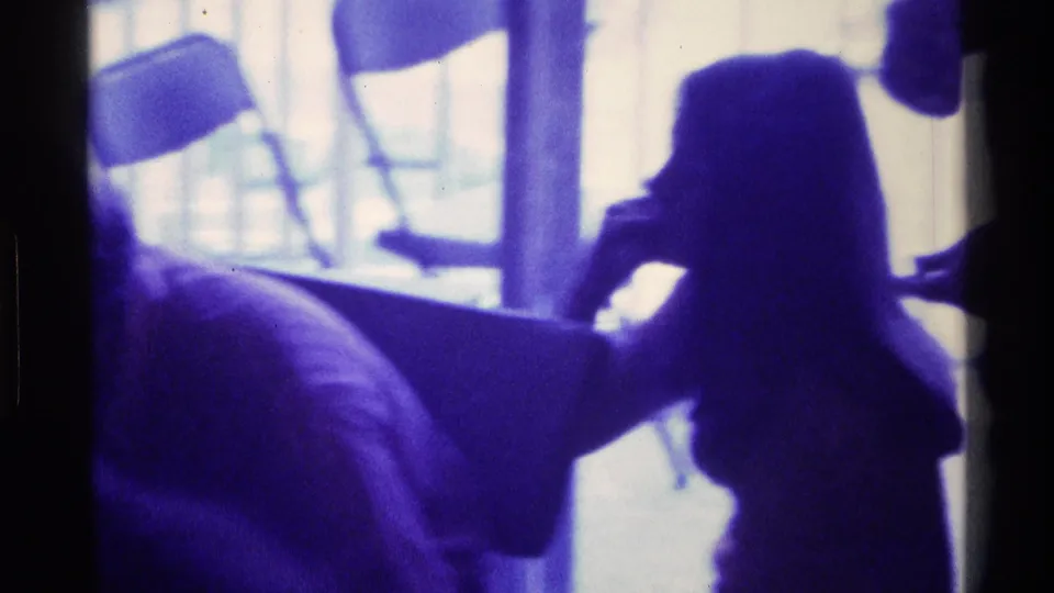 Home-movie frame from Kentucky in 1976 showing seated silhouetted people near a bright window and folding chairs.