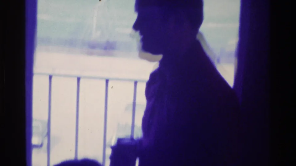 1976 Kentucky home-movie frame with a standing adult silhouette in profile near a bright window and railing.