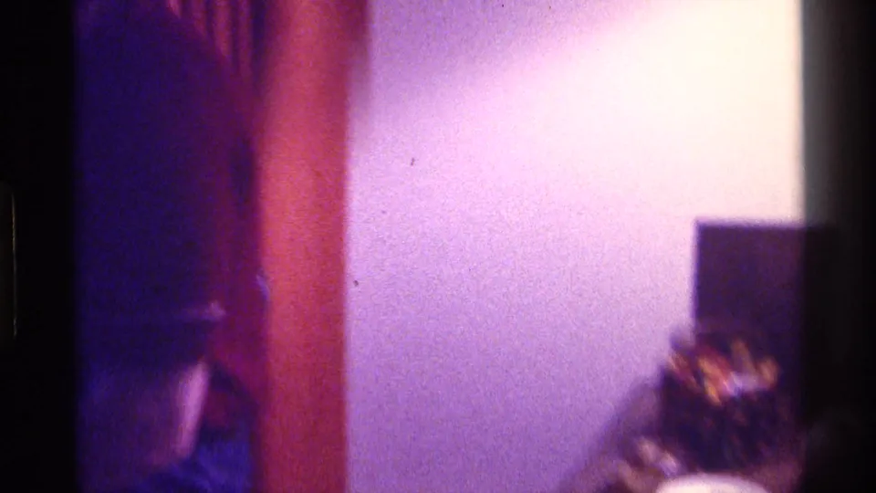 The camera pans across a dim 1976 Kentucky interior, showing warm wall color, shadowed shapes, and a partial person at the frame edge.