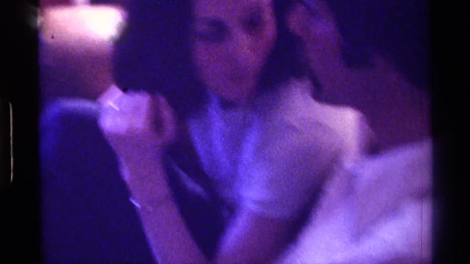 A seated woman and another adult appear in a low-light 1976 Kentucky home-movie frame with strong purple color shift.