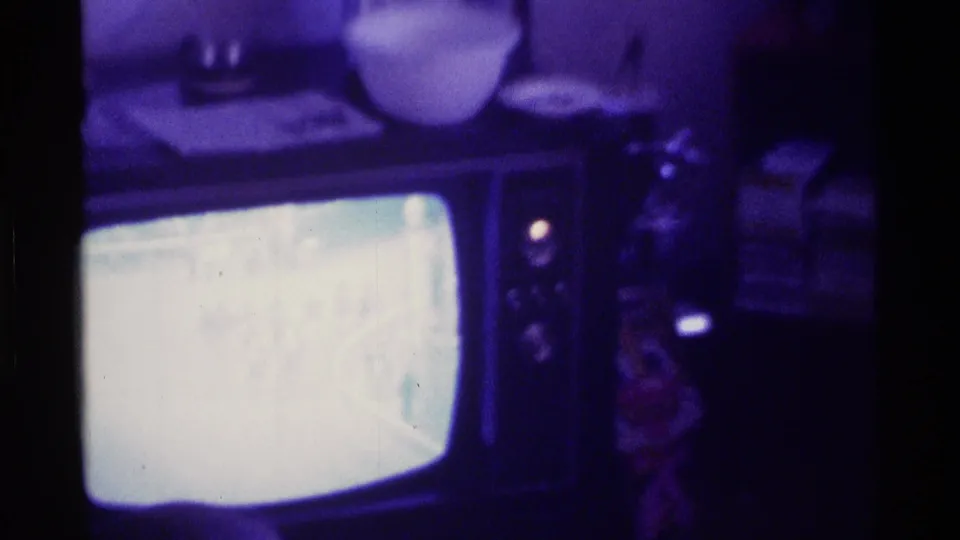 Purple-tinted 8mm frame of a CRT television in a Kentucky interior, with the racing broadcast washed out by screen brightness.