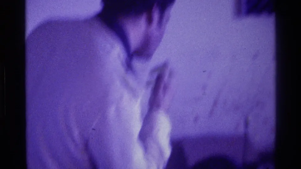 A viewer in a light shirt appears near the television area in this 1976 Kentucky home-movie frame, with facial detail obscured by blur and low light.