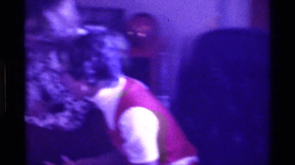 1976 Kentucky home-movie frame with a young person in a red and white shirt leaning near a table in a dim private room.