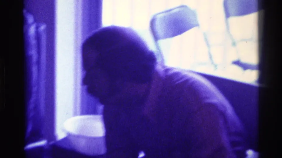 Purple-tinted 1976 Kentucky interior frame showing a seated adult leaning forward near a table, with folding chairs visible beyond.