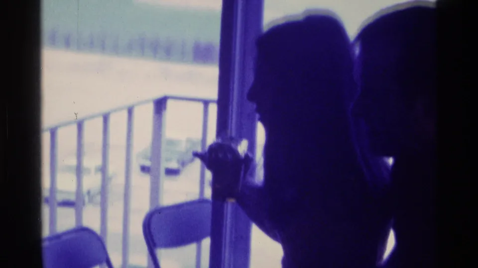 1976 Kentucky home-movie frame of a silhouetted person near a doorway, with railing and folding chairs visible through the bright opening.