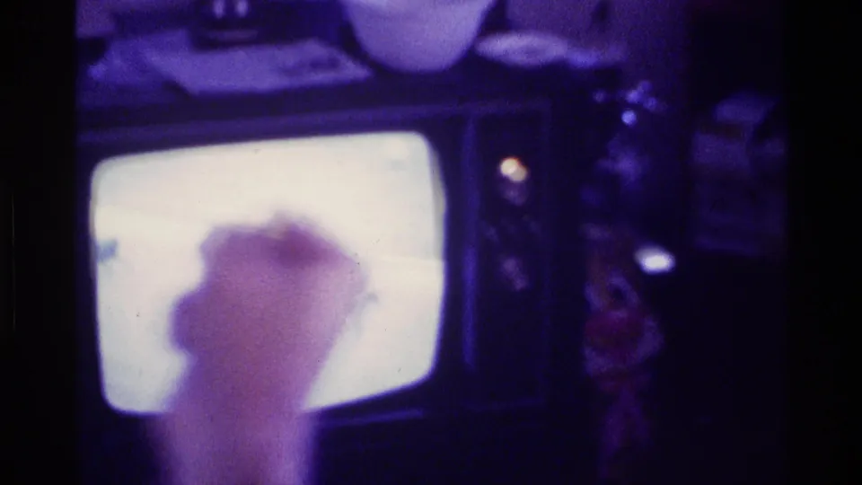 A hand or arm moves near the glowing television during a 1976 Kentucky home-movie view of family TV watching.