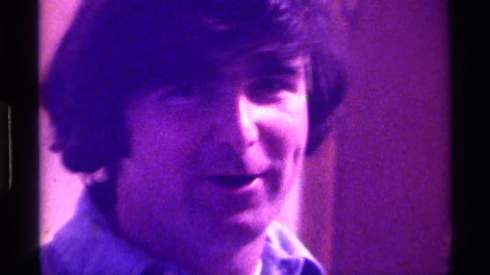 The young man appears to speak softly toward the camera in this purple-shifted 1976 Kentucky home-movie frame.