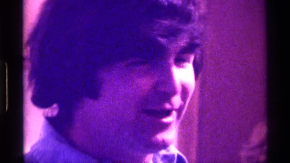 A close Super 8 frame shows the young man smiling faintly, with his face partly turned away in the indoor Kentucky scene.