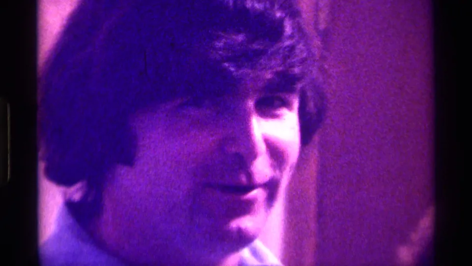 The young man remains in close-up, smiling slightly as the purple-tinted 8mm frame softens facial detail.