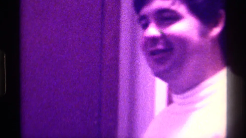 In the final frame, the young man smiles near an indoor wall, wearing a light turtleneck or high-collared top in purple-shifted 8mm footage.