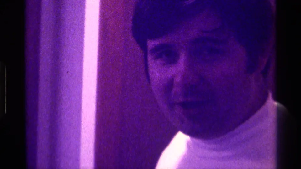 Young man in a white turtleneck faces the camera in purple-lit 1976 Kentucky home-movie footage.