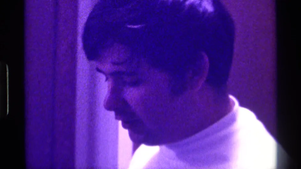 Side-profile close-up of the man in a white turtleneck during a purple-lit 1976 Kentucky home-movie performance.