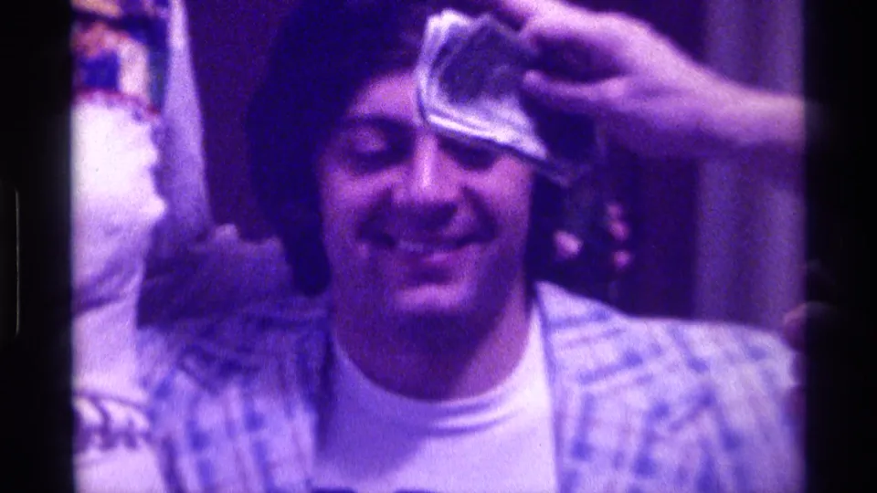 Young man smiles toward the camera while a hand holds a dollar bill against his forehead in 1976 Kentucky home-movie footage.