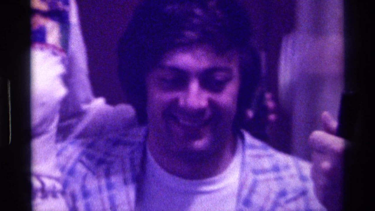 1976 Kentucky home-movie frame showing an adult man laughing in a dim interior, with another person's raised arm beside him.