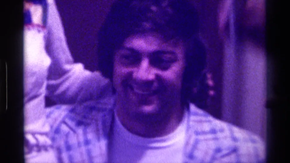 1976 Kentucky 8mm frame of a smiling adult man in a patterned shirt, filmed indoors with a raised arm visible at left.