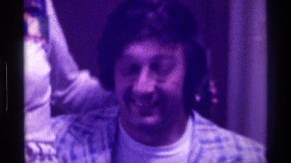 1976 Kentucky home-movie frame showing a man laughing openly indoors, with a nearby person's arm and shoulder partly in view.