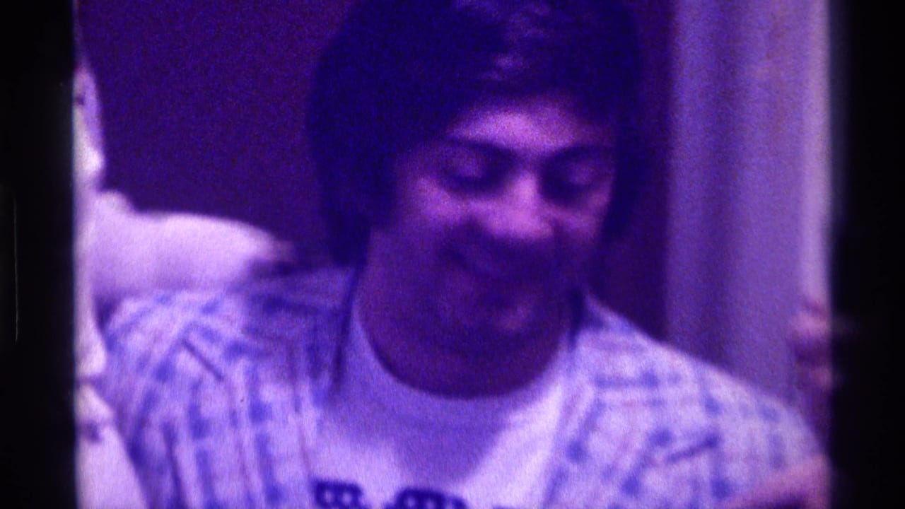 Young man in a plaid shirt smiles indoors under purple-tinted light.