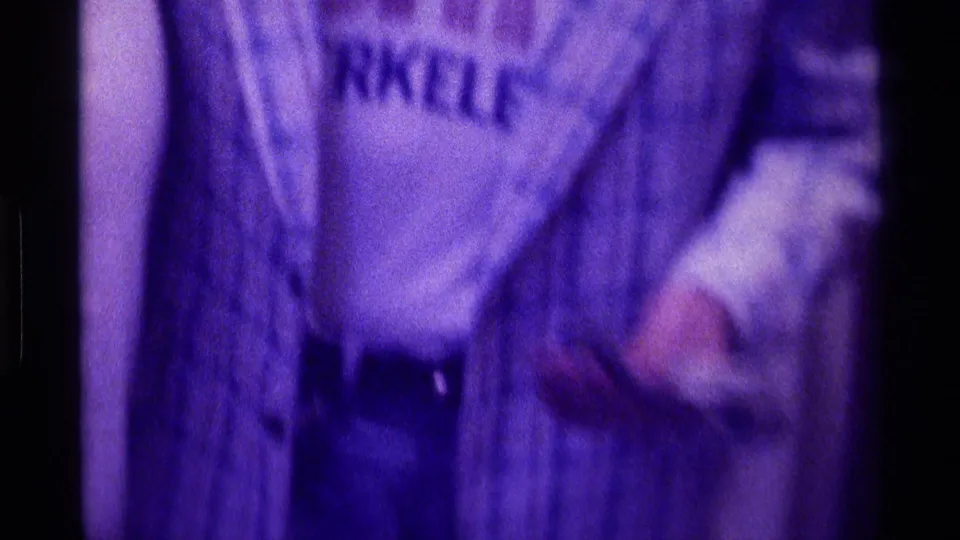 Close torso framing shows the plaid shirt, partial printed lettering on a white T-shirt, and a small object held near the waist.