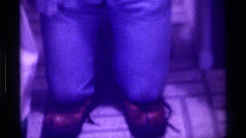 Lower-body framing shows fitted jeans, brown shoes, and tiled flooring in the purple-tinted 8mm home-movie shot.