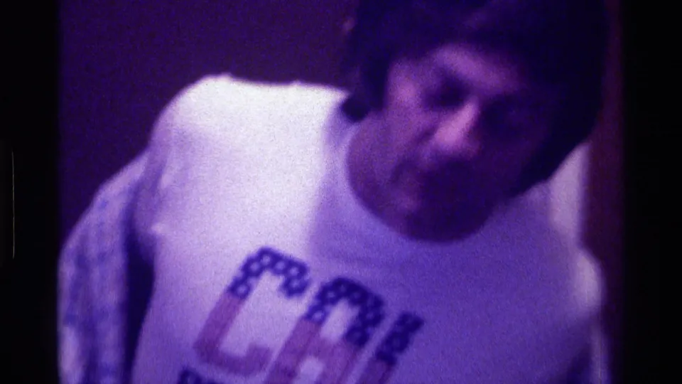 1976 Kentucky 8mm frame of a young man in a CAL BERKELEY T-shirt, filmed close-up in a dim interior.