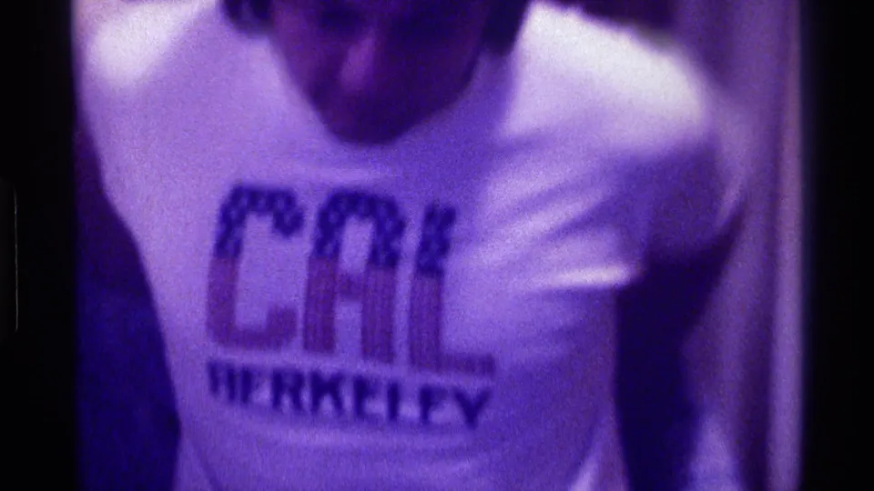1976 Kentucky home-movie frame of a man adjusting a white CAL BERKELEY T-shirt in a dim, purple-tinted interior.