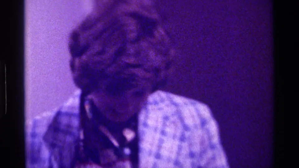 Kentucky 1976 8mm frame of a person in a patterned jacket looking downward in a dim, purple-tinted interior.