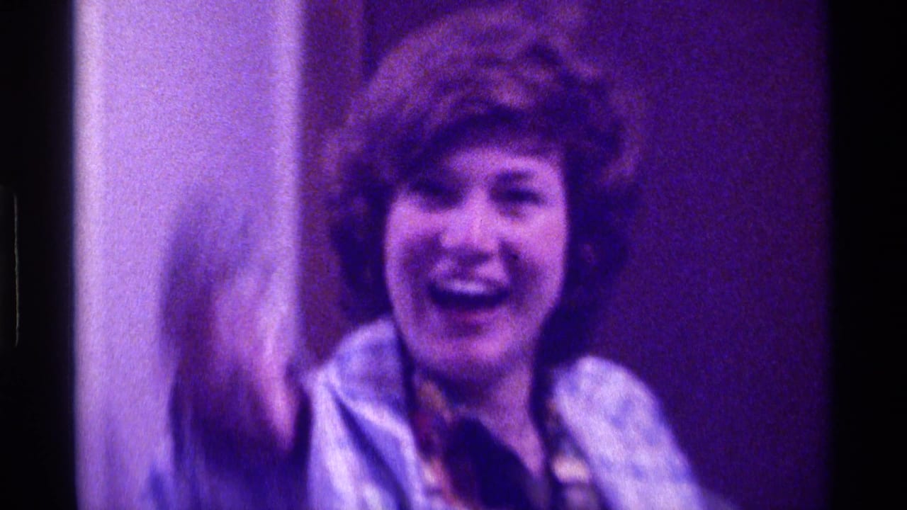 Woman in a blue plaid coat smiles and waves toward the camera indoors.