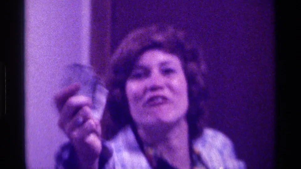 Adult woman in 1976 Kentucky footage raises folded money toward the camera, wearing a blue plaid coat and patterned scarf in a private interior.