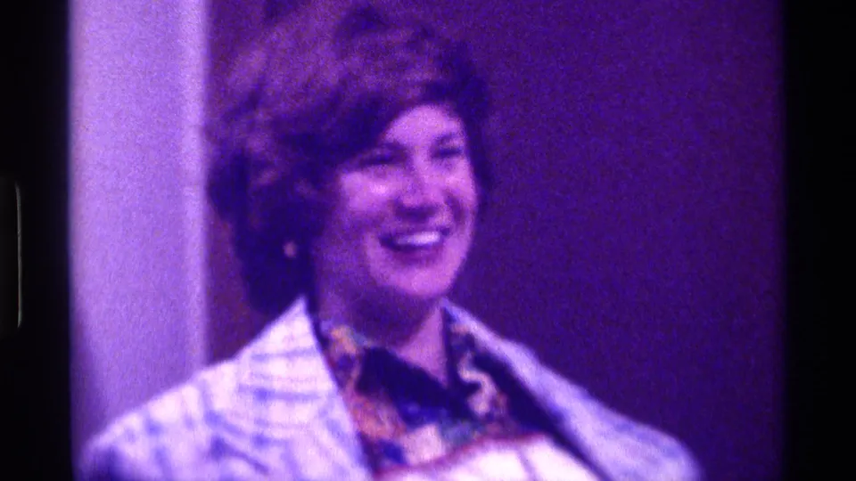 Adult woman in a blue plaid coat smiles to the side in grainy 1976 Kentucky home-movie footage with a patterned scarf visible.