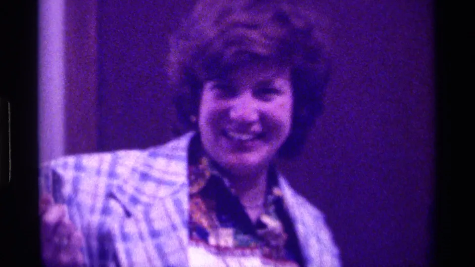Adult woman in 1976 Kentucky home-movie footage smiles toward the camera, with her blue plaid coat and patterned scarf visible under purple-tinted film color.