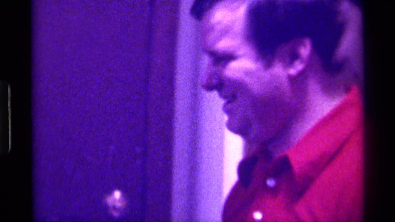 Adult man in a red polo shirt seen in profile beside a doorway under purple light.