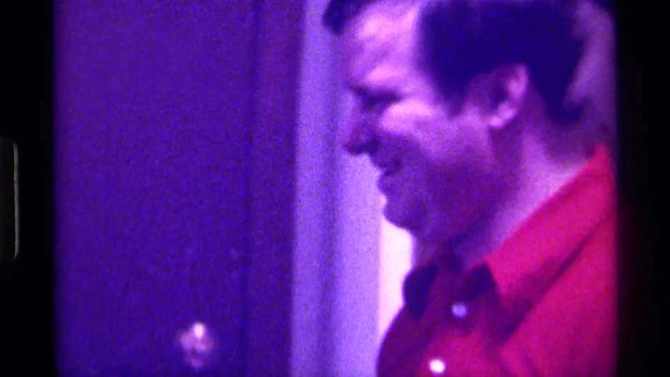 In 1976 Kentucky home-movie footage, an adult man in a red polo shirt stands in profile near a doorway under strong purple lighting.