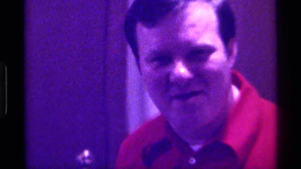 A man in a red polo shirt faces the camera in purple-tinted 1976 Kentucky 8mm footage, with a doorway visible beside him.