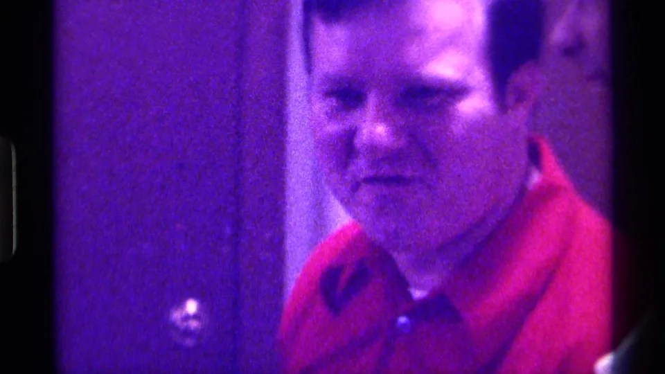 The man in a red polo shirt looks toward the camera in a purple-lit Kentucky home movie, with another partial face near the frame edge.