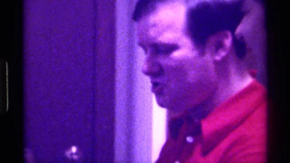 Adult man in a red shirt appears in profile in a Kentucky 1976 home-movie frame, with a doorway and dark interior behind him.