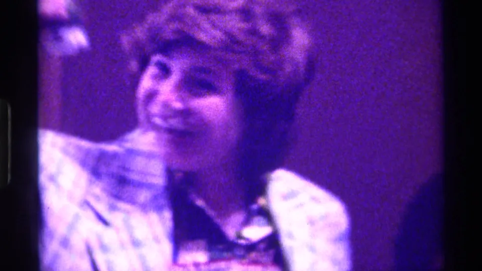 Smiling woman in a blue plaid jacket holds a gold-colored trophy in this Kentucky 1976 indoor home-movie frame.