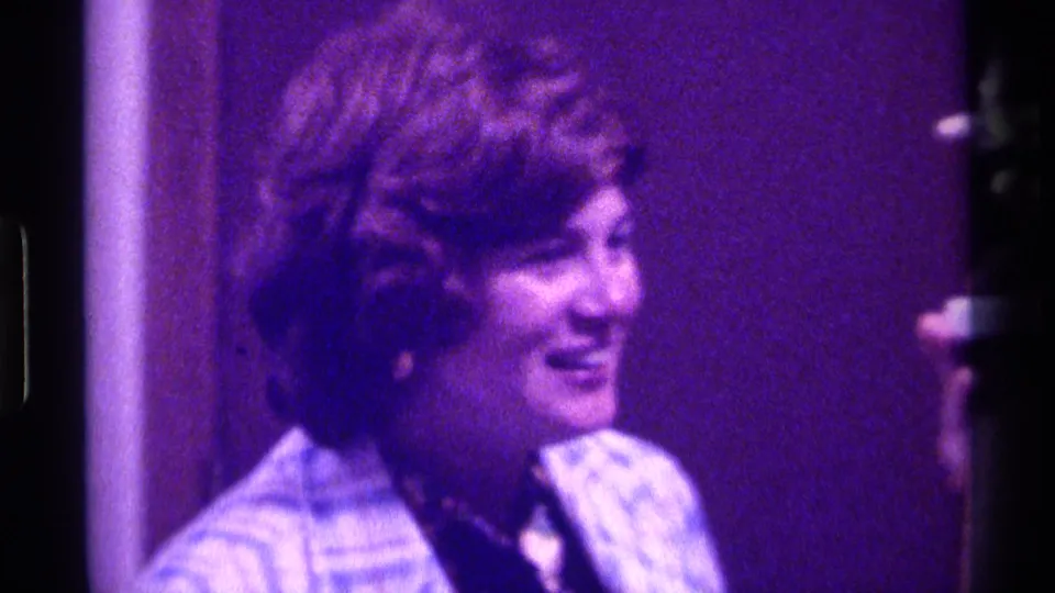 Woman in a blue plaid jacket smiles in profile inside a Kentucky 1976 home-movie scene, with the trophy partly near the frame edge.